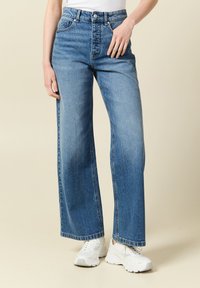 Jeans z wysokim stanem i szerokimi nogawkami w niebieskim kolorze, z przednimi kieszeniami i lekko wyblakłym wykończeniem, zestawione z białymi butami sportowymi.