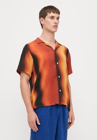 Camisa de botones de manga corta con un patrón abstracto en naranja, negro y amarillo. Fabricada en un tejido suave, presenta un cuello y un corte relajado.