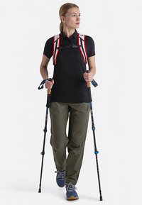 Polo noir, pantalon cargo vert olive, bretelles de sac à dos rouges et blanches, et bâtons de randonnée bleus avec poignées en bois. Pose de marche sur fond blanc.