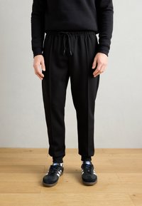 Black jogger pants with an elastic waistband and drawstring; features side seams and ribbed cuffs. Worn with black sneakers and blue accents.