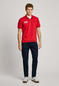 Red polo shirt with a collar and short sleeves, featuring a Union Jack patch. Paired with navy blue trousers and white sneakers.