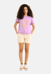 T-shirt en coton violet clair avec du texte graphique jaune et un design de palmier, assorti à un short couleur crème et des baskets blanches avec des accents jaunes.