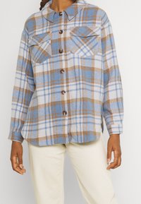 Camicia a maniche lunghe con bottoni, in pattern a quadri blu, bianco e marrone, indossata con pantaloni crema, che mostra il torso e le braccia senza testa.