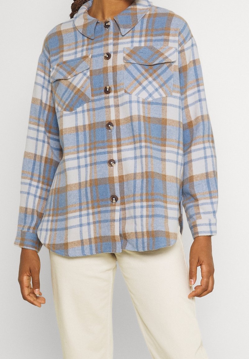 Camicia a maniche lunghe con bottoni, in pattern a quadri blu, bianco e marrone, indossata con pantaloni crema, che mostra il torso e le braccia senza testa.