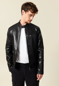 Giubbotto di pelle nera con colletto alto, tasche con zip e dettagli trapuntati sulle spalle. Indossato sopra una maglietta bianca e pantaloni neri.