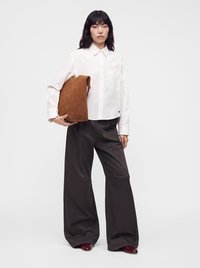 Femme portant une chemise blanche à boutons et un pantalon large noir tenant un grand sac marron sur fond blanc uni.
