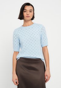 Pull en tricot bleu clair à manches courtes avec un motif en losange et des bords festonnés, associé à une jupe en satin marron.