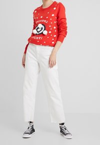 Pull rouge à pois blancs avec le texte « SNOWBALL FIGHT ! », orné de pompons blancs ; associé à un jean blanc large et des baskets montantes noires.