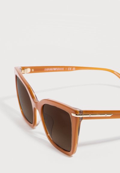 Καφέ διαφανή γυαλιά ηλίου με τετράγωνο πλαίσιο, σκοτεινούς φακούς και λεπτές χρυσές λεπτομέρειες στους βραχίονες, απεικονιζόμενα σε λευκό φόντο.