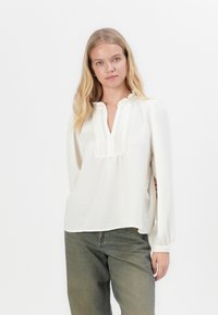 Blouse blanche avec une surface texturée, encolure froncée et un décolleté en V sans boutons. Manches longues avec poignets élastiques, portée avec un pantalon vert.