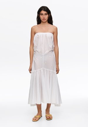 Mujer con cabello largo y oscuro lleva un vestido midi blanco sin tirantes con pequeños adornos blancos y sandalias amarillas, de pie sobre fondo blanco.