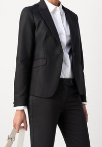 Blazer negro a medida con textura suave, cierre de un solo botón y solapa con muesca, combinado con una camisa blanca y pantalones negros.