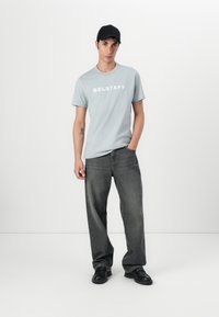 Világoskék póló fehér "BELSTAFF" felirattal, laza sötétszürke farmernadrággal és fekete cipővel párosítva. A modell fehér háttér előtt áll.