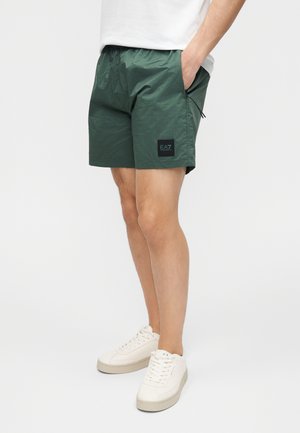 Homme portant un short à carreaux verts avec le logo EA7, des baskets blanches et un t-shirt blanc, debout les mains dans les poches devant un fond uni.