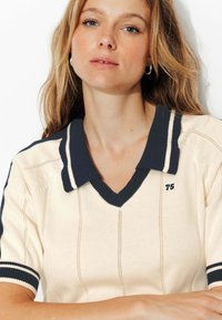 Polo en tricot beige avec col marine et manches rayées. Présente un col en V, des coutures verticales et une petite broderie "75".