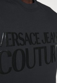 Schwarzer Pullover mit eingeprägtem Text "Versace Jeans Couture" auf der Brust, Nahaufnahme der Stofftextur und der Schriftzüge.