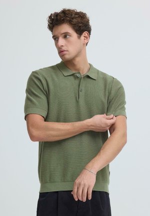 Mann med krøllete brunt hår som justerer ermet på en olivengrønn kortermet polo, mot en ensfarget lys bakgrunn.