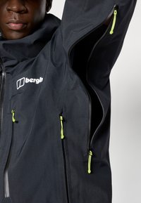 Zwarte waterdichte jas met een hoge kraag, ritszakken met felgroene trekbanden en een opvallend logo op de borst.