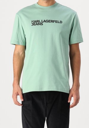 Hombre con camiseta de manga corta verde claro con texto negro "KARL LAGERFELD JEANS" en el pecho y pantalones negros.