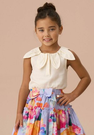 Fille aux cheveux foncés en chignon souriante, portant un haut sans manches crème avec un col en pétales et une jupe fleurie colorée avec un grand nœud.