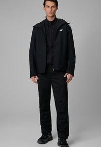 Homme debout devant un fond gris, portant une veste noire zippée, un pantalon noir et des baskets noires, face à l'avant avec les mains détendues sur les côtés.
