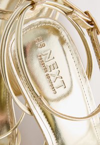 Sandale métallique dorée avec des brides, indiquant la taille 6,5/40 et le nom de la marque Next Forever Comfort sur la semelle intérieure.