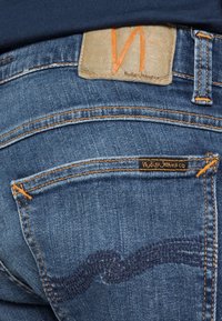 Mörkblå denimjeans med synlig orange sömnad, en texturerad läderetikett med präglad logotyp och en unik dekorativ design på bakfickan.