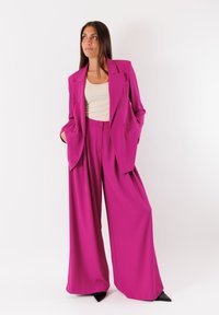 Blazer oversized fucsia con pantaloni a gamba larga abbinati, entrambi in un tessuto liscio. Presenta un colletto a rever e chiusura con bottoni.