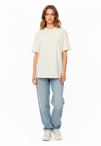 T-shirt oversize color crema con maniche corte, abbinato a jeans di denim blu chiaro e sneakers bianche. Design semplice e dal fit rilassato.