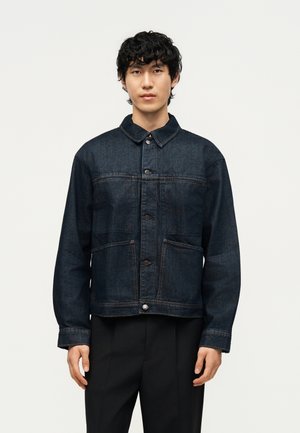 Jeune homme aux cheveux noirs bouclés portant une veste en jean foncée boutonnée et un pantalon noir sur un fond clair uni.