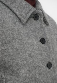 Szürke gyapjúkabát klasszikus gallérral, fekete gombokkal és texturált felülettel. Az anyag lágy és melegnek tűnik.
