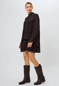 Vestido negro transparente con estampado de leopardo, cuello alto, mangas largas de campana y dobladillo en capas con volantes. La modelo lleva botas marrones hasta la rodilla con detalle de hebilla.