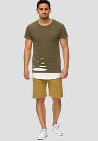 T-shirt en coton vert olive avec de grandes découpes, porté sur une chemise blanche, assorti à un short beige et des baskets blanches.