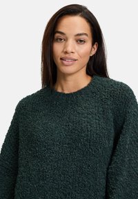 Dunkelgrüner strukturierter Pullover mit rundem Halsauschnitt, aus weichem, flauschigem Material. Das Design ist kastig und hat lange Ärmel.