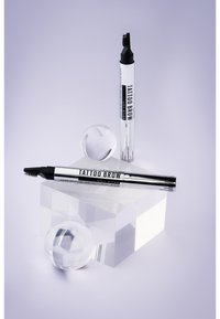 Tattoo Brow potlood en liftstick, zilver verpakt, met een schuine punt, textuurrijke borstel, gepositioneerd op heldere geometrische blokken en een kristallen bol.