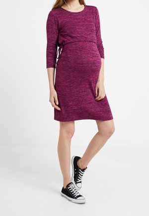 Pregnant woman wearing a fitted, long-sleeve, knee-length pink dress with black and white sneakers, standing against a plain white background.