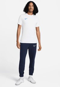 T-shirt bianco Nike con logo blu, abbinato a pantaloni jogger blu navy con logo bianco e polsini alla caviglia. Scarpe sportive bianche.
