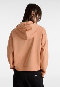 Sudadera corta de color melocotón con capucha ajustable, hecha de una tela suave. Pantalones negros con una etiqueta de logo visible en el bolsillo trasero.