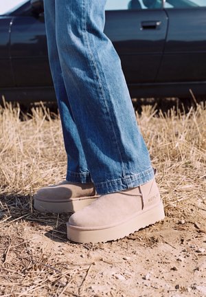 Une personne portant un jean bleu et des chaussures beiges à plateforme se tient sur de l’herbe sèche à côté d’une voiture sombre.