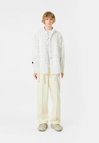 Camisa blanca de gran tamaño con diseño de patchwork; combinada con pantalones crema y zapatos grises texturizados. Fondo liso.