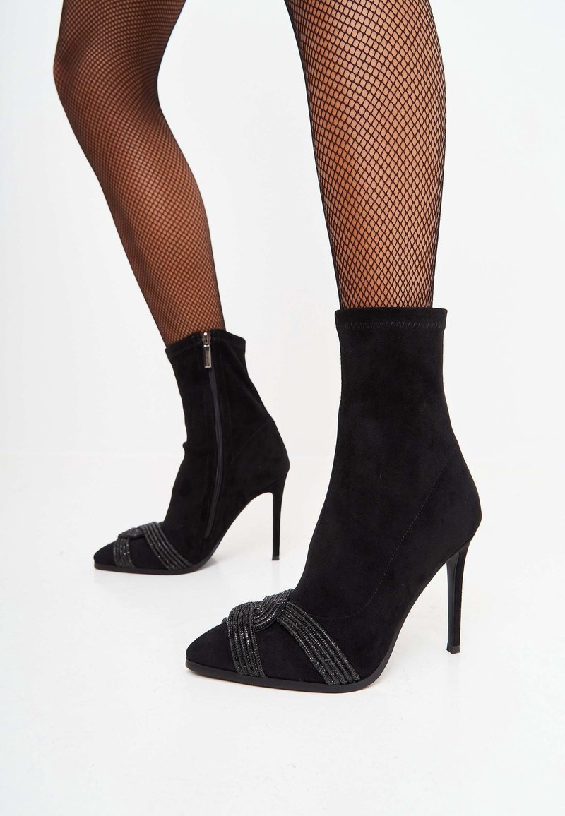 Bottines en suede noir avec un bout pointu et un talon aiguille, dotées d'une sangle décorative nouée et d'une fermeture éclair sur le côté pour un enfilage facile.
