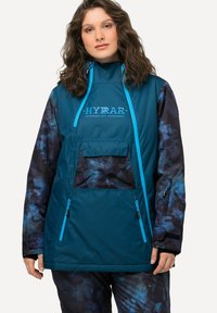 Giacca esterna color teal con colletto alto, accenti blu e maniche con pattern astratto blu scuro. Presenta una tasca frontale e tasche laterali con zip.