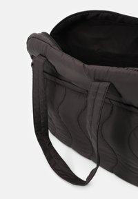 Černá prošívaná taška s hladkým povrchem, dvěma ušima a zipovým otvorem, který odhaluje černý interiér. Obsahuje zakřivený šev.