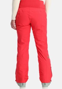Helle rote Schneehosen mit einem gerippten hohen Bund, Reißverschlüssen an den Knöcheln und Branding am unteren Bein. Hergestellt aus wasserabweisendem Material.