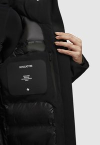Sort jakke med en puffer inderside, der har en lynlås-lomme og et label med teksten "KHUJOTM." Texturen er glat med en mat finish.