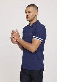 Polo bleu marine en coton, doté d'un col classique, de deux boutons et d'accents rayés blancs sur les manches courtes. Détail du logo rouge.