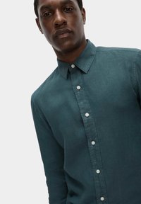 Camisa de lino color verde azulado con cuello puntiagudo, que presenta botones blancos en la parte delantera y un acabado texturizado. Diseño de corte casual.