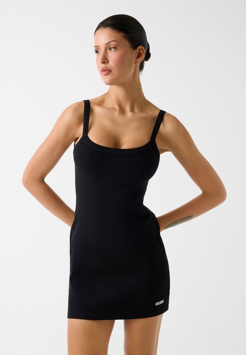 Vrouw met donker haar in een lage knot, gekleed in een nauwsluitende zwarte mouwloze jurk met dunne bandjes en een klein wit Guess-logo vlak bij de zoom.
