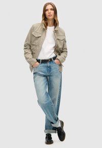 Veste en toile gris clair avec plusieurs poches, t-shirt blanc, jean bleu clair à coupe décontractée et chaussures noires avec une finition texturée.