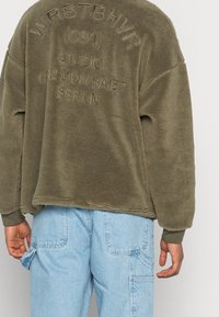 Felpa verde in pile con testo in rilievo sulla schiena, caratterizzata da una vestibilità ampia e polsini a coste, abbinata a pantaloni cargo azzurri.